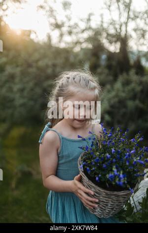 Potrait eines kleinen Mädchens, das sich um Blumen im Garten kümmert, lila Blumenglocken im Topf hält. Stockfoto