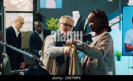 Ein afroamerikanischer Mitarbeiter im Einzelhandel zeigt eine neue Modekollektion im Kaufhaus des Einkaufszentrums. Schwangere Assistentin macht Farbvorschläge an Stammkunden, Konsumverhalten. Kamera A.. Stockfoto
