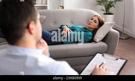 Eine junge Frau liegt auf einer Couch beim Termin eines Psychologen. Ein brauner, männlicher Psychologe mit Brille führt eine psychologische Sitzung durch Stockfoto