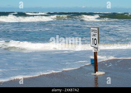 Es gibt eine Geschwindigkeitsbegrenzung von 10 Schildern und ein paralleles Parkschild an einem Holzpfosten an einem Sandstrand. Es gibt einen stürmischen Strand mit Wellen und Strömungen an der Küste. Stockfoto