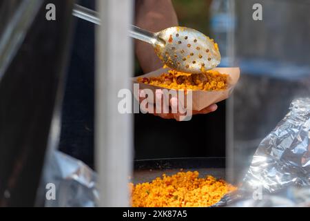 Ein Straßenverkäufer löffelt eine Schüssel mit scharfem indischem Reis. Der Metalllöffel hat kleine Löcher. Die quadratische Schüssel besteht aus Pappe. Stockfoto