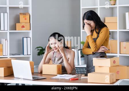 Zwei stressige Geschäftsfrauen verwalten Online-Bestellungen in einem modernen Büro mit Kartonschachteln und Laptop Stockfoto