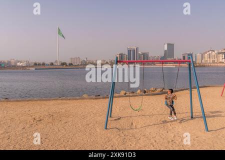 Der arabische Junge auf Schaukeln an der Küste des Hafens von Jeddah mit dem Stadtpanorama, der riesigen saudischen Flagge, den Wolkenkratzern. Stockfoto