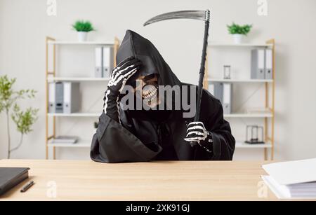 Müder Mann im Halloween-Sensenmann-Todeskostüm, der Sense hält, die am Schreibtisch im Büro sitzt Stockfoto