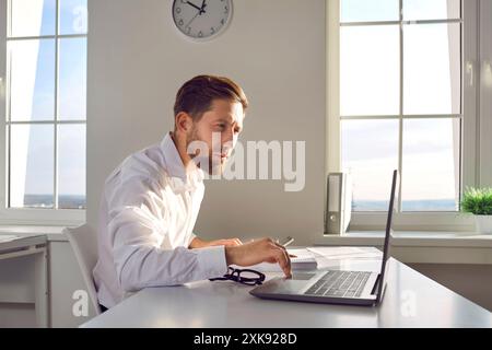 Junger Geschäftsmann, der am Schreibtisch an seinem Arbeitsplatz im Büro an einem Laptop arbeitet. Stockfoto