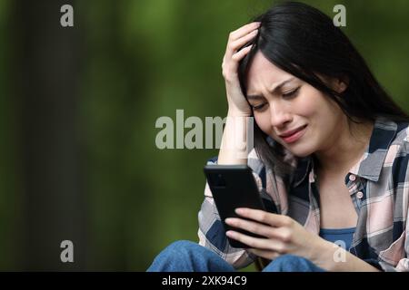 Besorgte asiatische Frau, die schlechte Nachrichten auf dem Handy überprüft und sich in einem Park beschwert Stockfoto