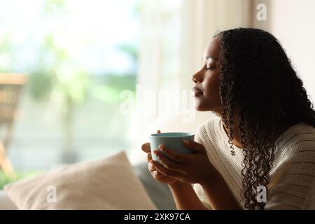 Seitenporträt einer schwarzen Frau zu Hause, die Kaffee trinkt und sich mit geschlossenen Augen entspannt Stockfoto