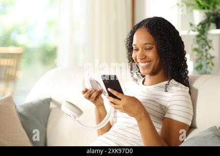 Glückliche schwarze Frau, die einen kabellosen Kopfhörer mit einem Smartphone verbindet, das zu Hause auf einer Couch sitzt Stockfoto