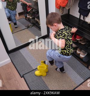 Ein Kind zieht Schuhe an, die im Flur stehen. Das Baby zieht gelbe Stiefel an, um spazieren zu gehen. Kind im Alter von 3 Jahren (dreijähriger Junge) Stockfoto