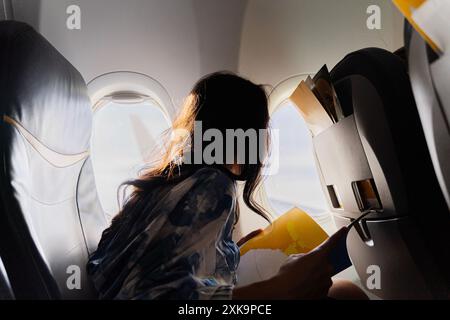 Eine schöne Asiatin liest im Flugzeug eine Zeitschrift. Eine junge Frau mit Buch, die aus dem Flugzeugfenster schaut. Freizeitreisen junge Passagierin liest bo Stockfoto