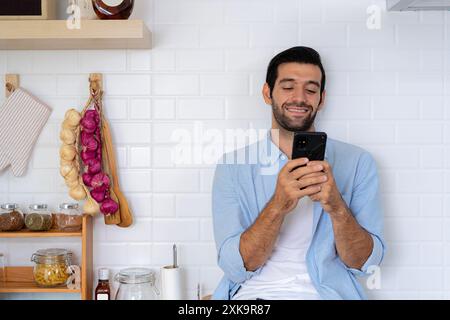 Fröhlicher bärtiger junger Mann, der auf dem Handy surft, in sozialen Medien surft und mit glücklichem Lächeln arbeitet, während er in der Küche zu Hause sitzt Stockfoto