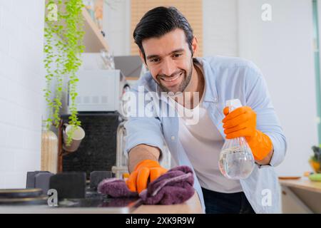 Reinige deine Sorgen weg. Ein junger Mann, der zu Hause eine Küchenzeile desinfiziert. Junger, gutaussehender Mann mit Schürze und Schutzhandschuhen, der Waschbecken und Wasserhahn abwischt Stockfoto