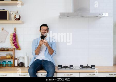 Schöner junger Mann glücklich, während er in der Küche zu Hause sitzt, am guten Morgen im gemütlichen Urlaub. Fröhlicher junger bärtiger Mann, der über Smartphone-Uin spricht Stockfoto