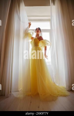 Die Silhouette einer Frau in einem goldenen, luxuriösen Kleid vor dem Hintergrund eines Fensters hält einen Vorhang mit den Händen. Elegante Dame in gelber Seide Stockfoto