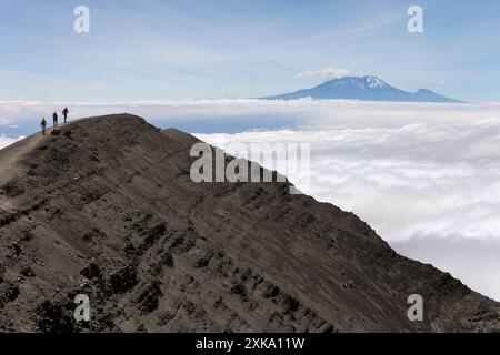 Drei Wanderer laufen auf einem felsigen Bergrücken des Mount Meru, mit dem Kilimandscharo im Hintergrund. Stockfoto