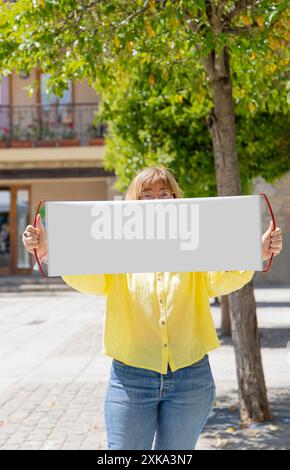 Eine Frau hält ein leeres Schild vor ihrem Gesicht. Das Schild ist weiß und hat keine Aufschrift. Die Frau trägt ein gelbes Hemd und blaue Jeans. Stockfoto