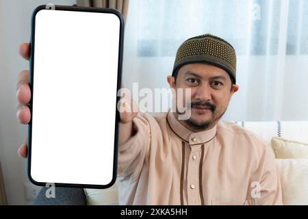 Junger arabischer Kerl, der neben einem großen Handy sitzt, mit leerem weißen Bildschirm, mit Mobilgerät, Mockup. Lächelnder junger arabischer Mann, der ein riesiges Smartphone demonstriert Stockfoto