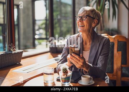 Reife Frau sitzt im Café und entspannt sich. Sie trinkt Kaffee und sendet telefonisch Nachrichten. Stockfoto