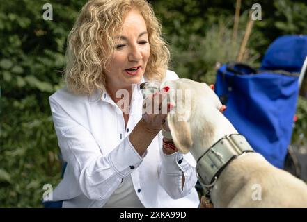 Reife Frau mit lockigem Haar, die liebevoll einen windhund während eines Picknicks streichelt, mit einem blauen Rucksack im Hintergrund und umgeben von Grün. Stockfoto