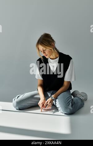 Die junge blonde Frau im stilvollen, legeren Outfit sitzt auf dem Boden mit den Händen in den Taschen, tief in Gedanken. Stockfoto