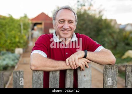 Ein erwachsener Mann, der entspannt neben dem Zaun im Garten steht und lächelt Stockfoto