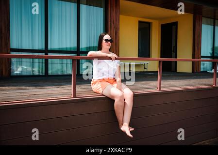 36 Jahre alte russin in orange Shorts und Sonnenbrille, die auf einem Balkon sitzt und am Sommertag genießt Stockfoto