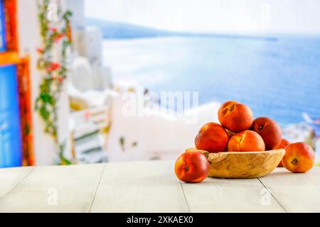 Frisches Obst von Pfirsichen in Holzschale auf Holzplatte mit wunderschönem verschwommenem Meer und blauem sonnigem Himmel Hintergrund mit einigen verschwommenen Gebäuden. Stockfoto