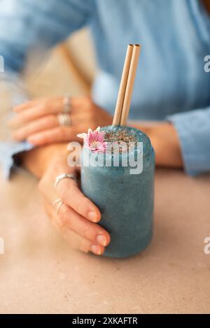 Blauer Spirulina veganer Smoothie mit Chiasamen, dekoriert mit essbaren Blüten. Frau, die ein Glas veganen Smoothie hält. Stockfoto