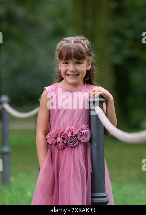 Porträt eines süßen 6-jährigen Mädchens mit langen dunklen Haaren in einem wunderschönen rosa Kleid. Ein Kind steht in der Nähe eines Zauns aus Seilen. Sie legte ihre Hände auf die Stockfoto