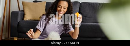 Eine junge Frau entspannt sich auf einer Couch und hält ein Stück Spielzeug, während ihr bichon Friseurhund neben ihr sitzt. Stockfoto