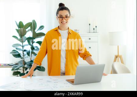 Porträt einer erfolgreichen, selbstbewussten jungen Frau aus arabischer oder indischer Herkunft mit Brille, Unternehmens-seo, Kreativmanager, IT-Experte steht neben ihrem Schreibtisch, blickt auf die Kamera und lächelt Stockfoto