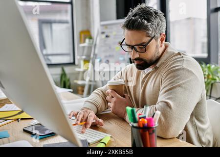 Ein gutaussehender Geschäftsmann mit Bart sitzt an seinem Schreibtisch in einem modernen Büro und tippt auf einer Computertastatur, während er eine Tasse Kaffee hält. Stockfoto