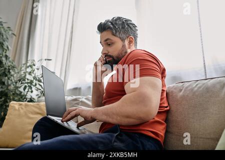 Ein bärtiger Mann in lässiger Kleidung arbeitet von zu Hause aus und multitaskt mit seinem Laptop und Handy. Stockfoto