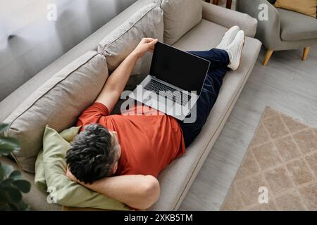 Ein bärtiger Mann in lässiger Kleidung entspannt sich auf einer Couch zu Hause und arbeitet ferngesteuert an seinem Laptop. Stockfoto