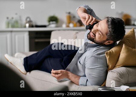 Ein bärtiger Mann in lässiger Kleidung entspannt sich auf einer Couch zu Hause und nimmt einen Anruf an. Stockfoto