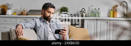 Ein bärtiger Mann in lässiger Kleidung entspannt sich auf einer Couch, benutzt sein Handy in einer Wohnung. Stockfoto