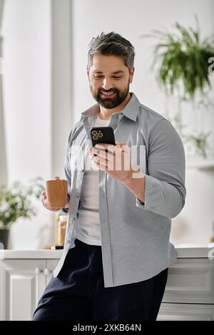 Ein bärtiger Mann in lässiger Kleidung steht in seinem Haus, lächelt, während er sein Handy in der Hand hält, während er eine Tasse Kaffee hält. Stockfoto