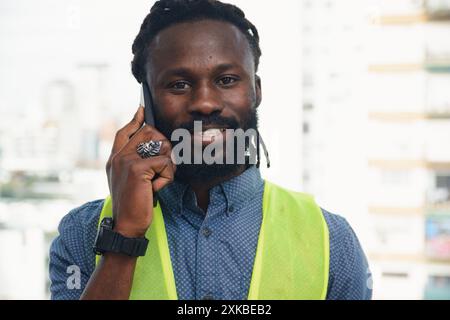 Ein glücklicher junger Mann afrikanischer Herkunft mit Bart und Dreadlocks spricht am Telefon, sieht in die Kamera und lächelt, er trägt eine reflektierende Jacke Stockfoto