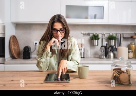 Lächelnde junge Frau, die Gläser trägt, Kaffee trinkt und einen Tablet-Computer benutzt, während sie in der Küche sitzt. Geschäftsfrau liest E-Mails morgens in ihr Stockfoto