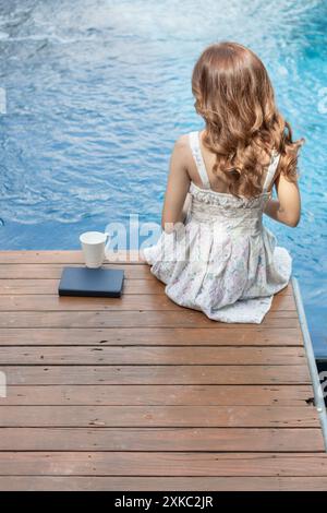 Eine junge Frau in einem alten weißen Kleid sitzt auf einer Terrasse am Pool und liest allein die Bibel, um Gottes Lehren aus der Bibel zu lernen und zu verstehen, die sie ist Stockfoto