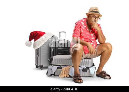 Trauriger reifer Mann, der zu weihnachten reist und auf einem vollgepackten Koffer mit unordentlichen Klamotten sitzt, isoliert auf weißem Hintergrund Stockfoto