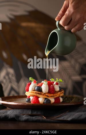 Ahornsirup wird über einen Stapel Pfannkuchen mit Erdbeeren und Beeren gegossen. Die Hand des Mannes gießen. Stockfoto