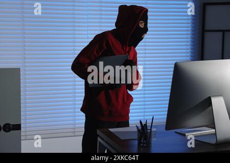 Dieb trägt Sturmhaube und stiehlt nachts im Büro einen Laptop. Einbruch Stockfoto