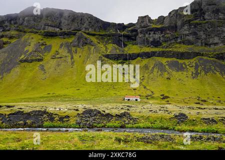 Verlassenes Haus auf dem Moos- und grasbedeckten grünen felsigen Berghang mit Wasserfällen und Fluss mit Schafen in Island während des bewölkten Sommers d Stockfoto