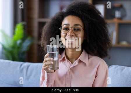 Porträt einer lächelnden Frau mit Brille, die zu Hause ein Glas Wasser hält. Förderung einer gesunden Lebensweise und Hydratation. Hintergrund mit gemütlichem Innenraum Stockfoto