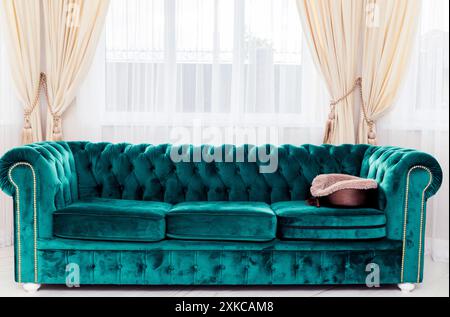 Ein luxuriöses grünes Samtsofa in einem hellen Wohnzimmer mit großen Fenstern und eleganten cremefarbenen Vorhängen. Stockfoto