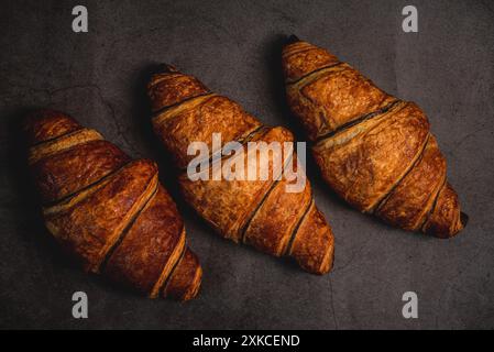 Drei frisch gebackene Croissants sitzen auf einer dunkelgrauen Arbeitsplatte, bereit zum Verzehr. Ihre goldbraune Kruste glitzert unter dem weichen Licht und lädt ein Stockfoto