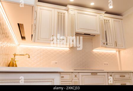 Luxuriöse weiße Küche mit goldfarbenen Schränken und moderner Beleuchtung. Ausgestattet mit gefliestem Backsplash, integrierten Geräten und Unterschrankbeleuchtung. Stockfoto