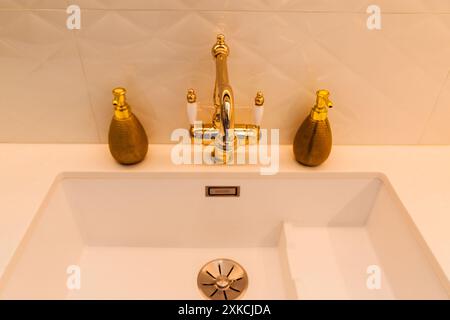 Blick von oben auf einen luxuriösen goldenen Wasserhahn mit passenden Seifenspendern auf einem weißen Küchenwaschbecken. LED-Beleuchtung unter Schränken verbessert das moderne Design. Stockfoto