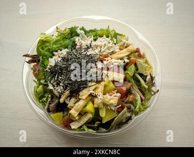 Poke Bowl hawaiianisches Meeresfrüchtegericht in einem auf dem Tisch isolierten Take-Out-Behälter (Wonton-Chips, Nori, Algensalat, Kani-Krabben, eingelegter Daikon-Radtich, Lachs, Stockfoto